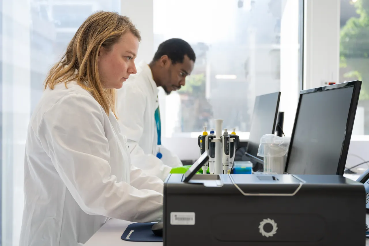 Picture of two scientist working in a lab, both on computers. A PromethION2 Integrated is in the foreground and pipettes are visible. © Alexandre Darmon / Art in Research