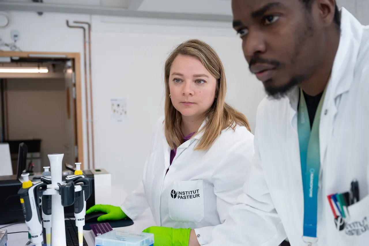 Two scientists are pictured in a lab. They are both wearing gloves and lab coats. They are both looking intently on something outside the picture. The female scientist in the back is holding a mouse and a keyboard is in view. © Alexandre Darmon / Art in Research