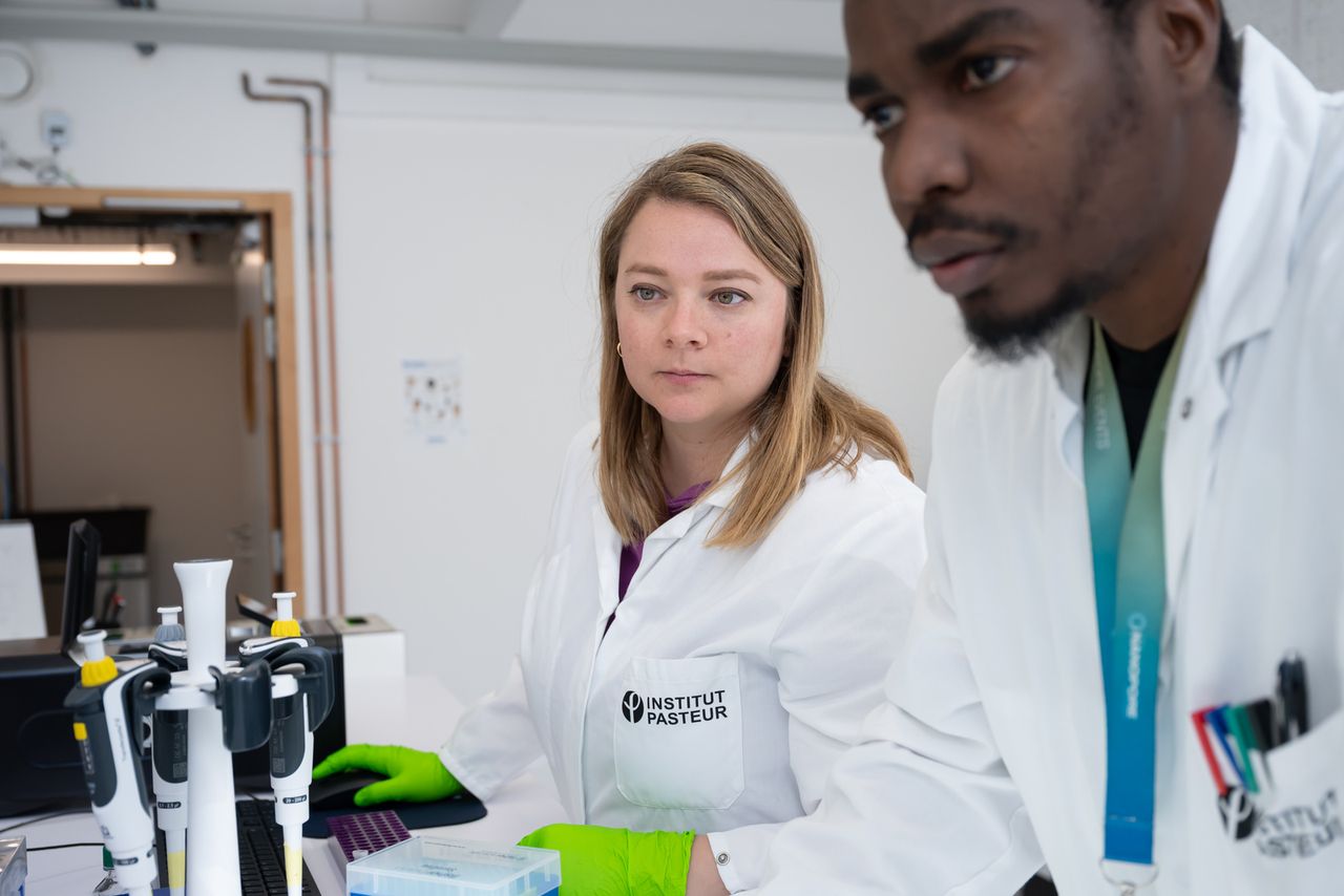 Two scientists are pictured in a lab. They are both wearing gloves and lab coats. They are both looking intently on something outside the picture. The female scientist in the back is holding a mouse and a keyboard is in view. © Alexandre Darmon / Art in Research
