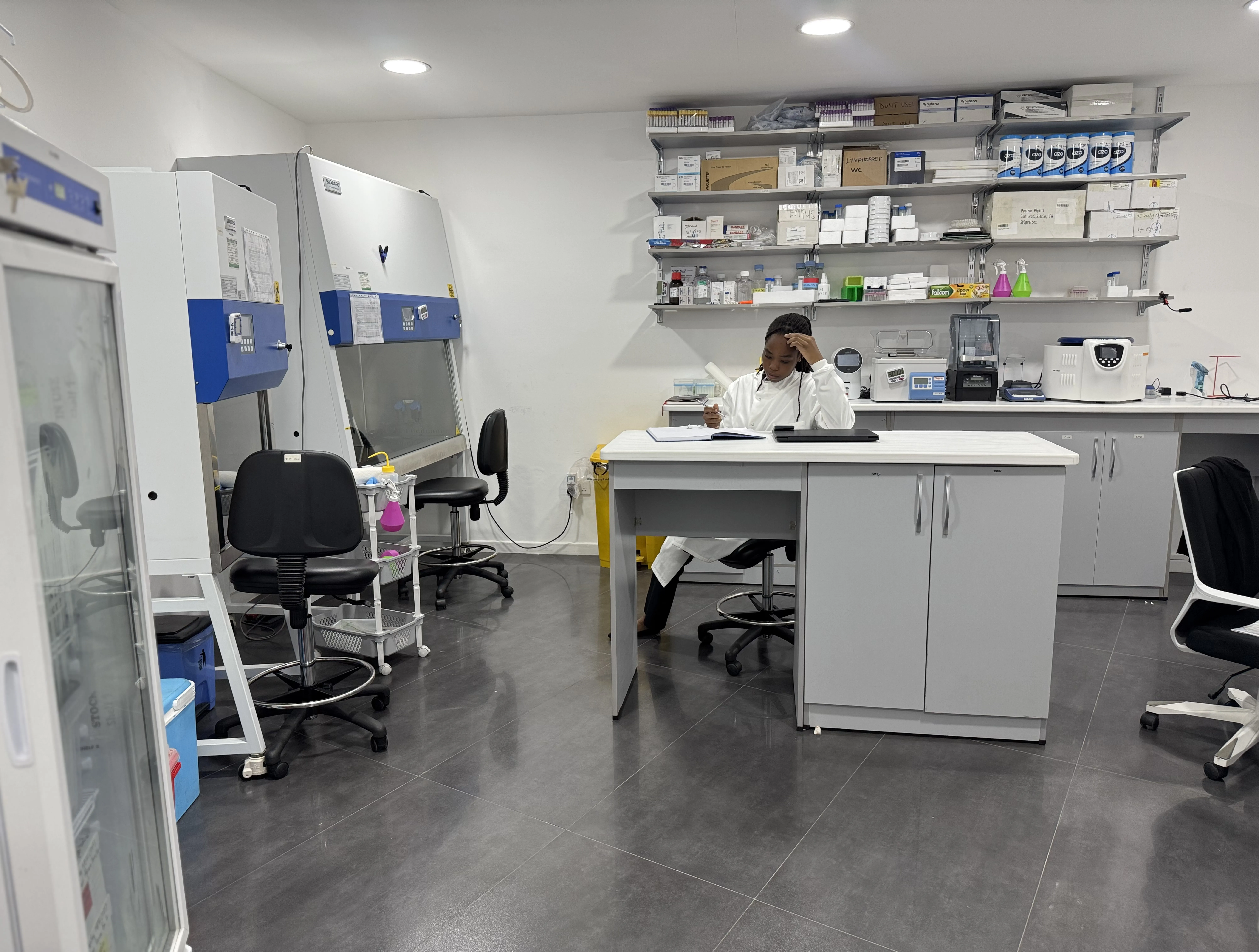 A woman in a lab coat is sitting on a white lab at a desk in the middle of a room, thinking whilst looking at a notebook