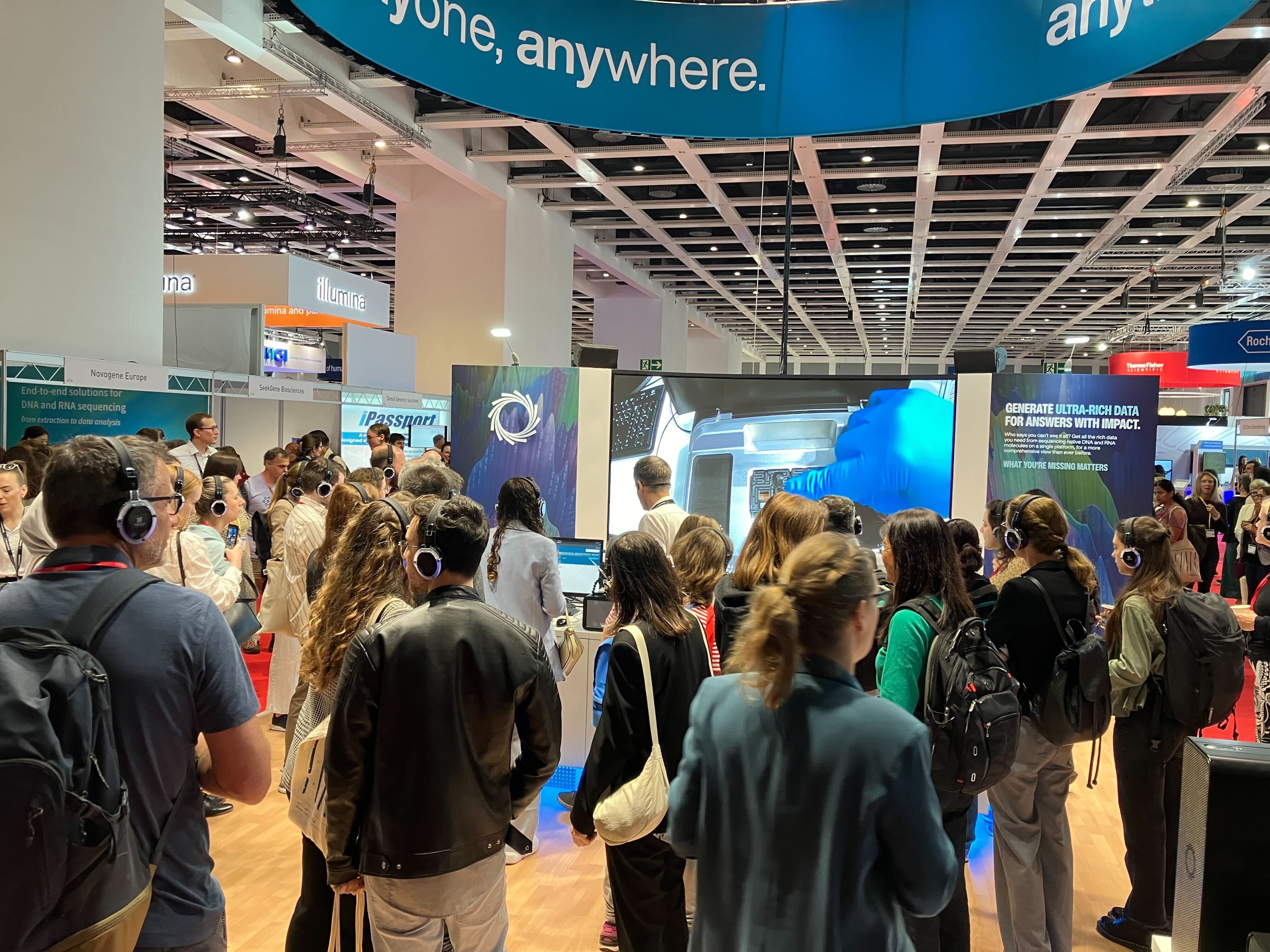 Lots of people crowded into the Oxford Nanopore booth, with a screen in the background which shows a hand adding a flow cell into a device