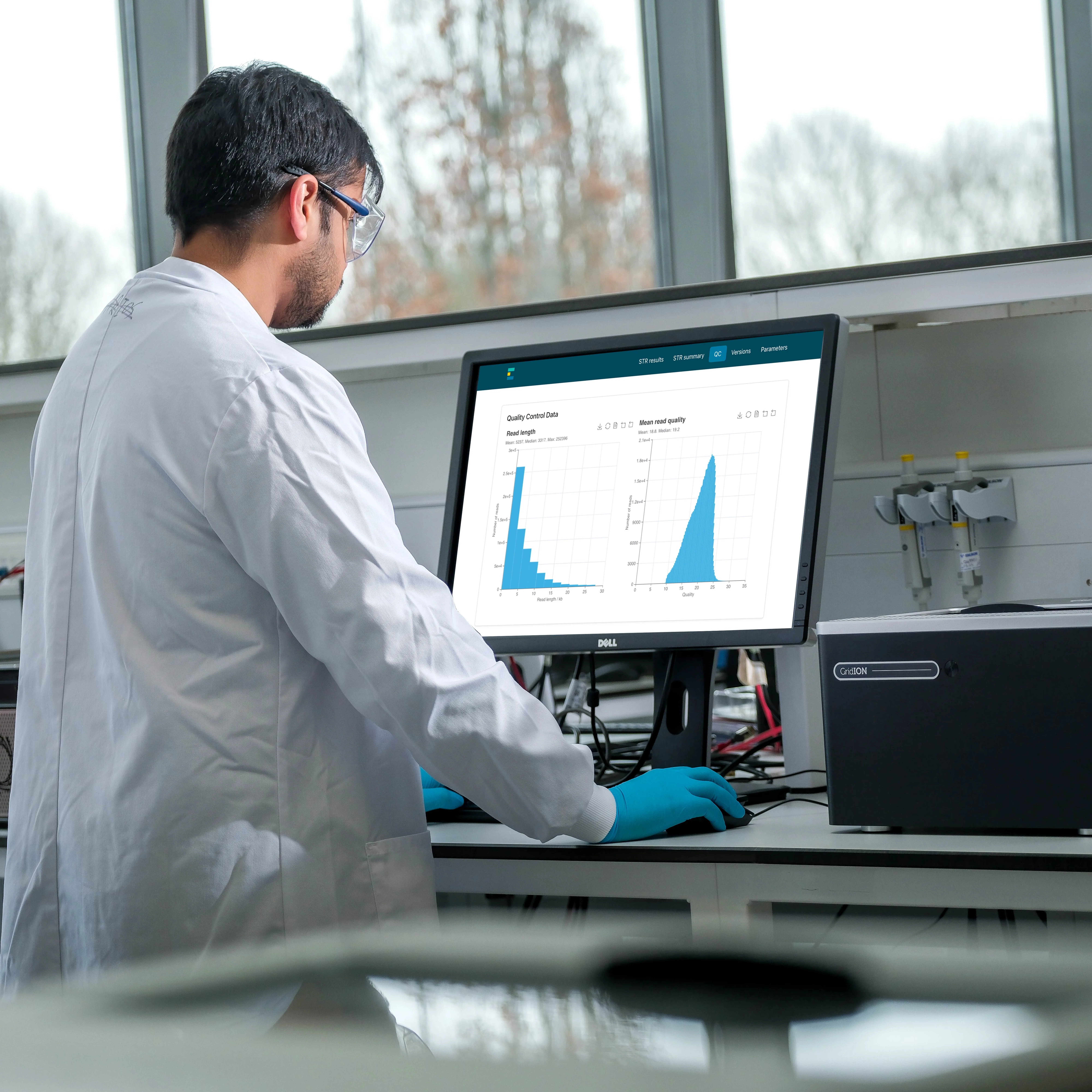 A Scientist wearing a white lab coat, goggles and gloves is on our computer looking at data within EPI2ME. They are in a lab and there are Oxford Nanopore sequencing devices to either side of them.